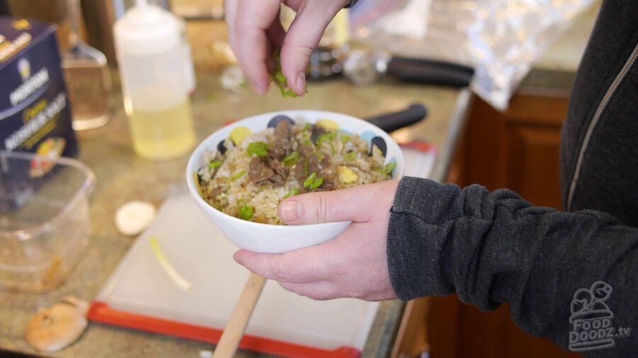 A bowl of leftover gyudonburi beef fried rice is held in one hand while the other adds slices of green onion to the top. It looks amazing and tastes even better!
