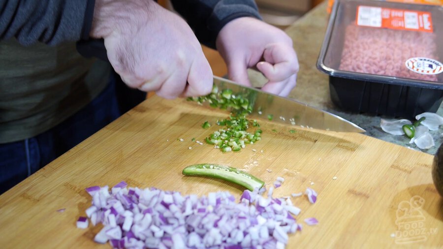 Chopped red onion sits in the foreground while serrano pepper is chopped on wood cutting board