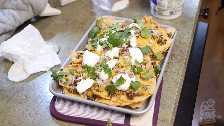 Baked nachos sheet is removed from oven and rests on baking mitts. A layer of torn cilantro is added on top along with dollops of sour cream.