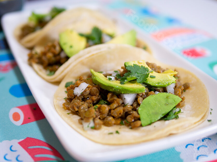 Three scrumptious homemade vegan corn tortilla lentil tacos sit on rectangular plate topped with avocado slices and cilantro. A colorful placemat is seen under plate.