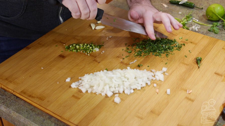 Cilantro is chopped with chef's knife on wood chopping board. Piles of chopped serrano pepper and onion can be seen.