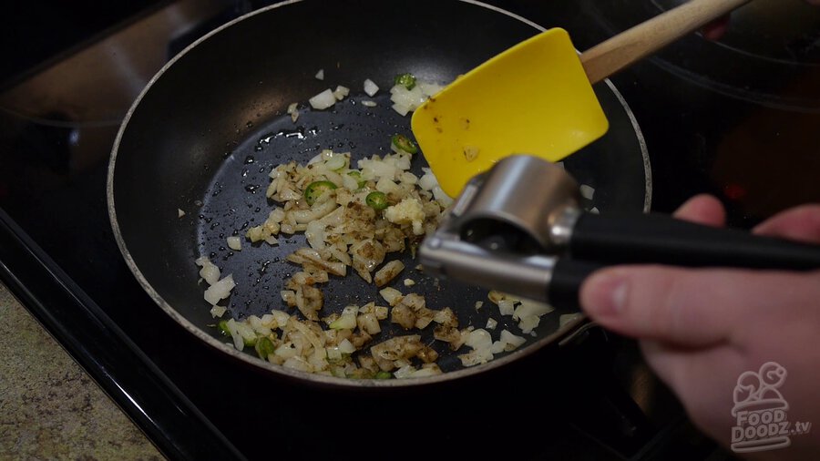 Garlic press is used to add minced garlic to skillet with chopped onion and sliced serrano peppers. A large spoon is used to spread garlic evenly.