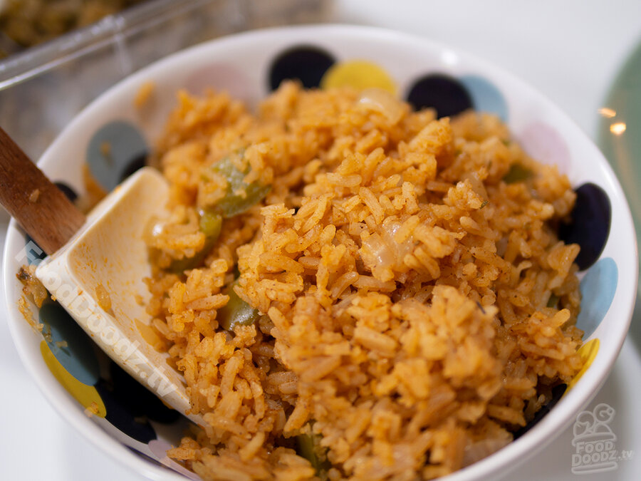 Tasty looking bowl of mexican rice with green bell peppers, onions, and tomatoes sits with large serving spoon to one side.