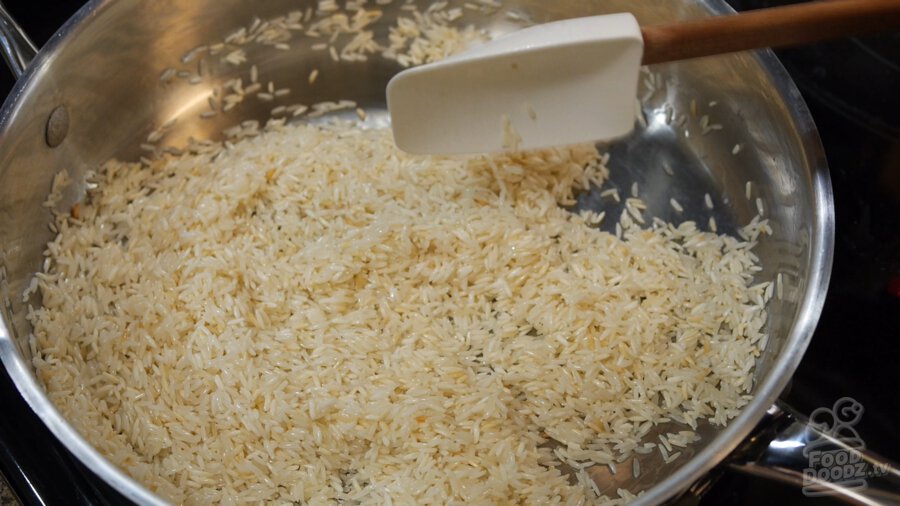 White rice toasts in oiled steel pan being stirred by large spoon