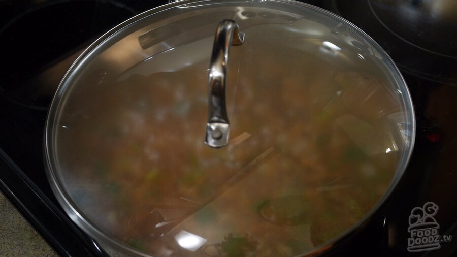 A lid is placed on top of pan and steam forms inside obscuring view of tomato sauce, vegetable broth, green bell pepper, onion, and toasted rice mixture inside