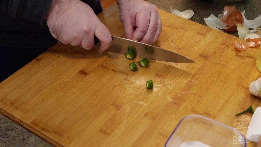 Slicing serrano peppers into half inch wide chunks with chef's knife on wood cutting board