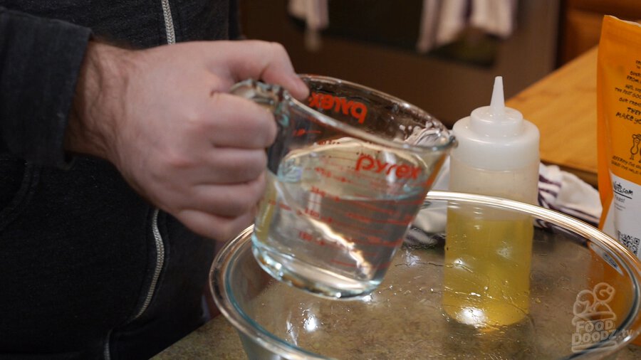 Water being poured out of glass measuring cup into large glass bowl sitting on digital scale