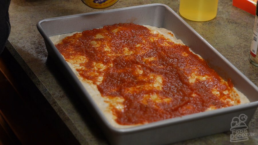 Sheet pan full of pizza dough sits covered in layer of tomato sauce
