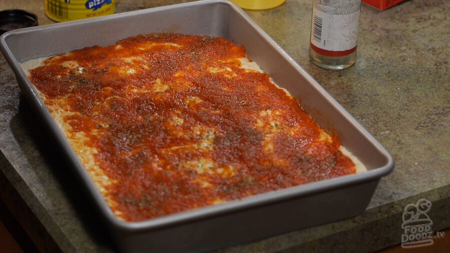 Sheet pan full of pizza dough sits covered in layer of dried basil, oregano, and garlic powered over tomato sauce