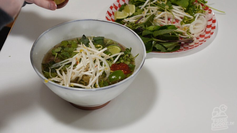 Completed Vietnamese Pho recipe in large bowl topped with beansprouts, cilantro, lime, chili paste, Thai basil, and sliced serrano peppers with garnish plate beside. Looks delicious! It is!