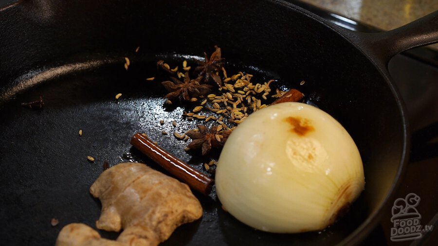 Star anise, cinnamon sticks, cloves, and fennel seeds added to charring onion and ginger halves in cast iron skillet