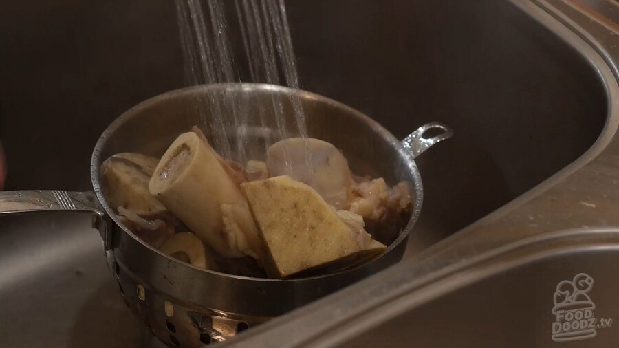 Boiled bones in colander being rinsed under faucet of sink