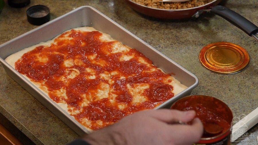 Sheet pan full of pizza dough sits covered in layer of crushed tomatoes while additional sauce spooned out of can