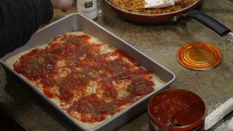 Hand sprinkles basil, oregano, and garlic powder on sauced pizza dough in sheet pan
