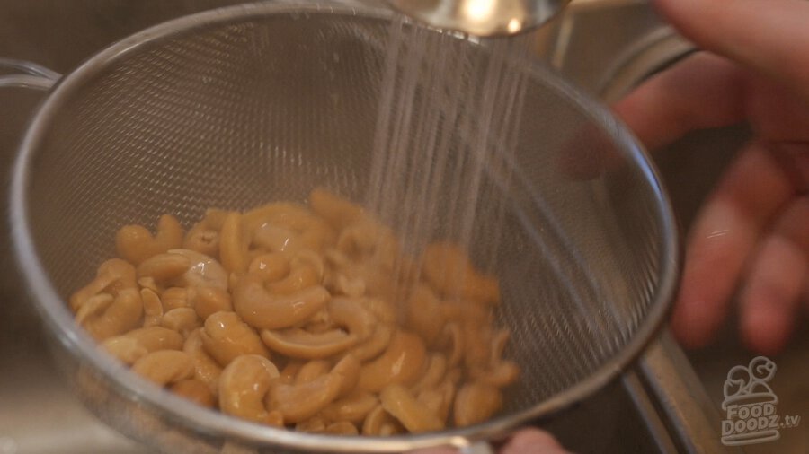 Rinsing boiled cashews in colander with faucet over sink