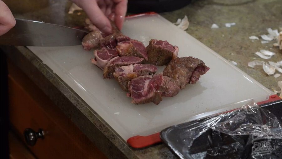 Slicing browned chuck roast beef into smaller cubes on cutting board with chefs knife