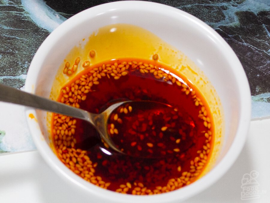 A deep red bowl of Chinese Sichuan Chili Oil sits with a spoon in it. Sesame seeds float on the surface.