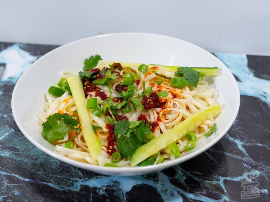 An extremely appetizing bowl of wheat noodles sit on a colorful faux marble place mat. Bright red chili oil, sliced scallions, cilantro, and cucumber sticks top the noodles.