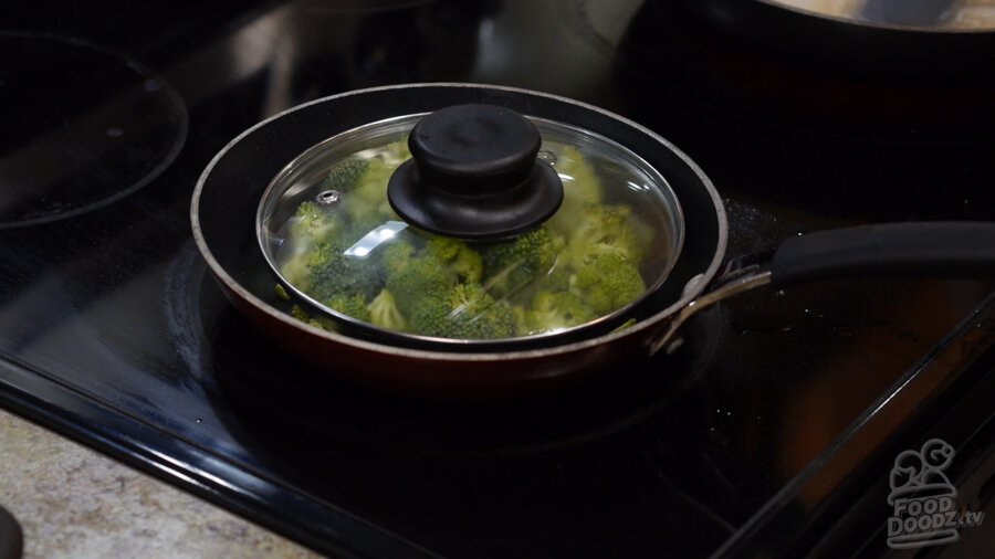Broccoli added to pan with a little splash of water and a lid placed on top