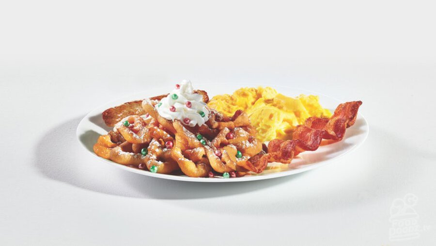 IHOP Little Elves Oh What Funnel Cakes Combo funnel cakes toped with powered sugar, glazed strawberries, whipped topping, and red and green elf sprinkles sits next to a breakfast plate with sausage, ham, bacon, hashbrowns, and eggs