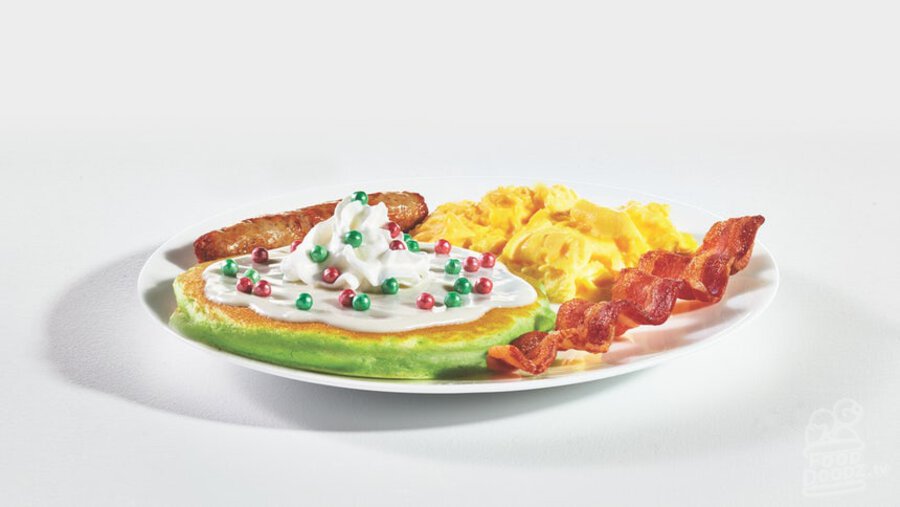 IHOP Little Elves Jolly Cakes Combo pancakes covered in sweet cream cheese white icing, whipped topping, and ornament sprinkles sits next to a breakfast plate with sausage, ham, bacon, hashbrowns, and eggs