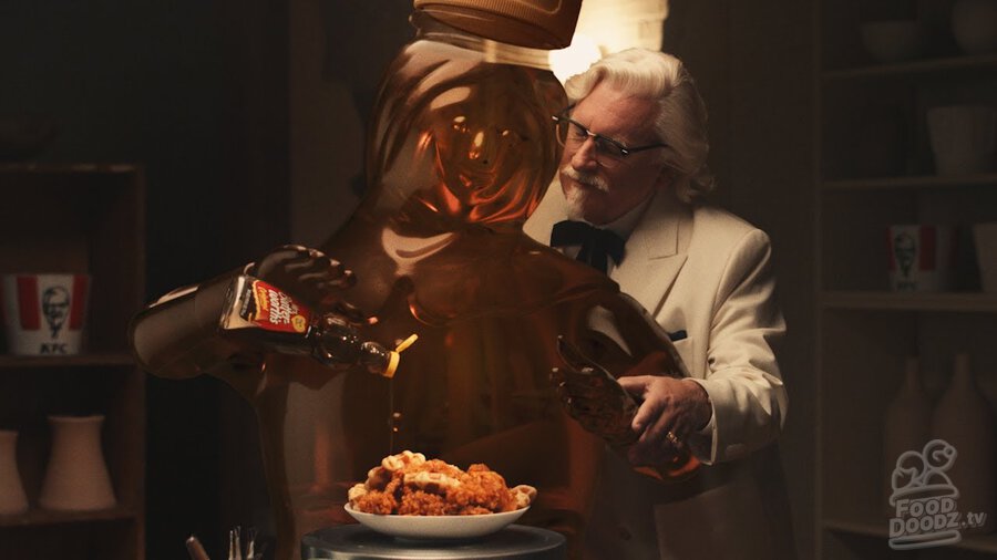 KFC (Kentucky Fried Chicken) Colonel Sanders stands behind Mrs. Butterworth while she pours syrup on a plate of chicken and waffles